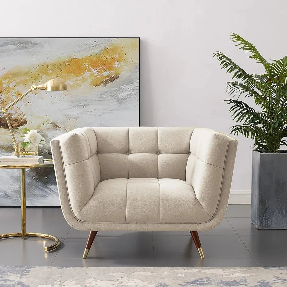 Mid-century modern armchair in light beige fabric with square tufting, curved back, and wooden legs with gold caps, next to a gold task lamp and a large plant.