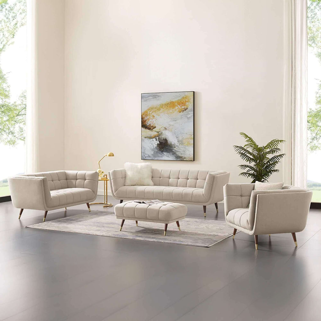 A full living room furniture set in a modern, brightly lit space, featuring a light beige tufted three-seater sofa with a white pillow, two matching armchairs, and an ottoman, arranged on a patterned rug under abstract art.