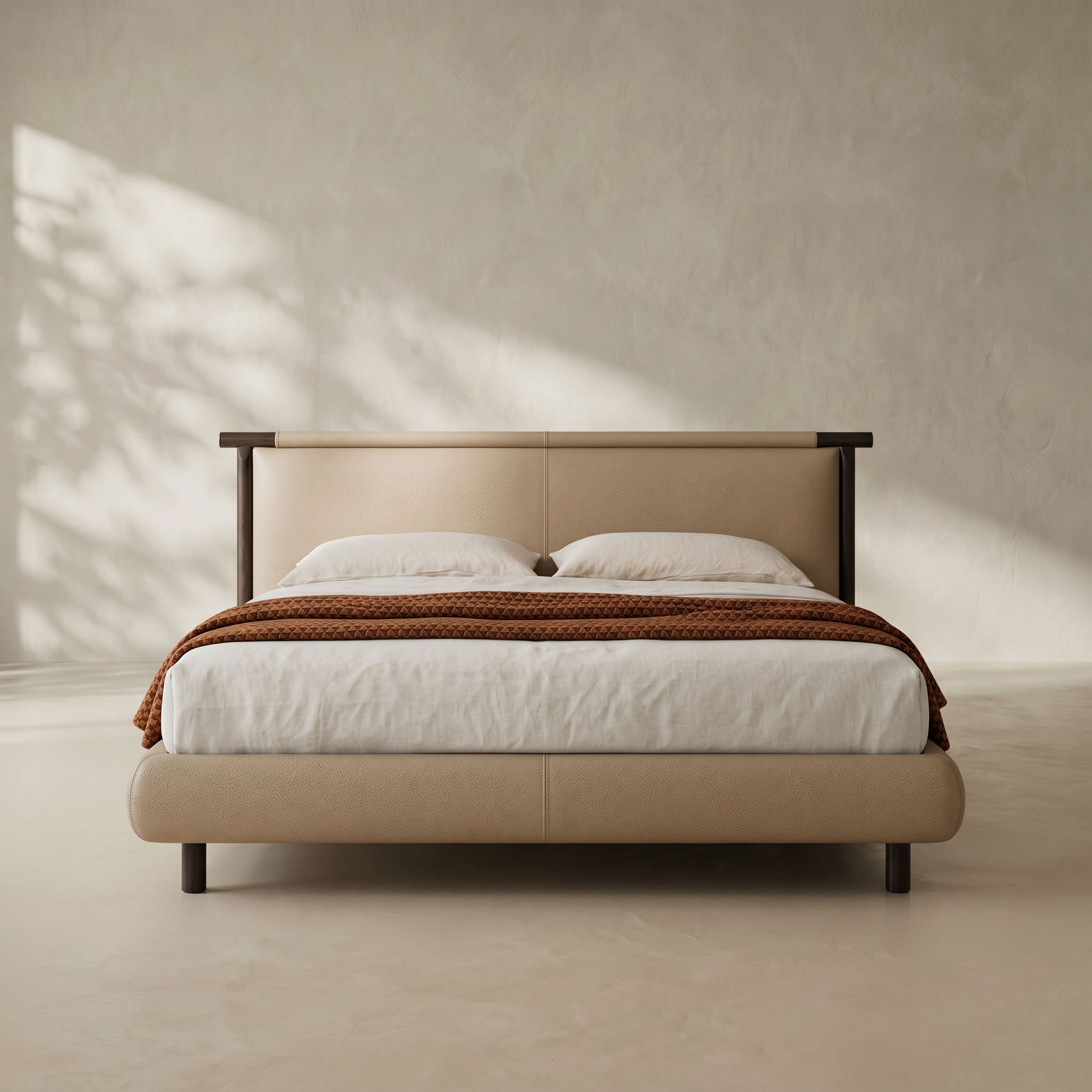 A modern upholstered bed with a beige leather-like frame and headboard, styled with white bedding and a brown patterned throw blanket, set against a textured beige wall with soft leaf shadows.