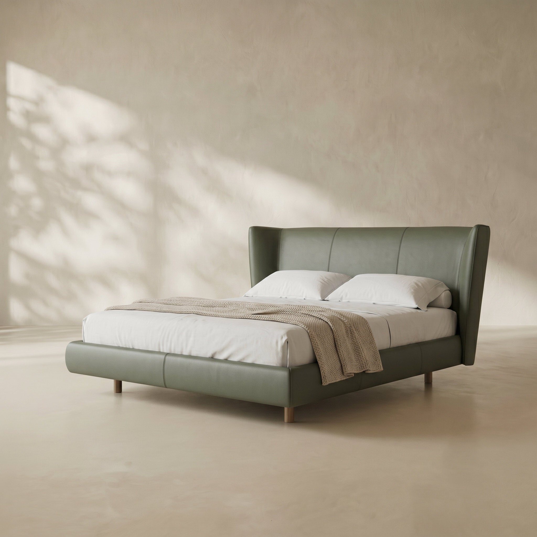 A modern platform bed with a tall wingback headboard upholstered in muted sage green leather, styled with white bedding and a beige throw blanket, set against a textured beige wall with soft leaf shadows.