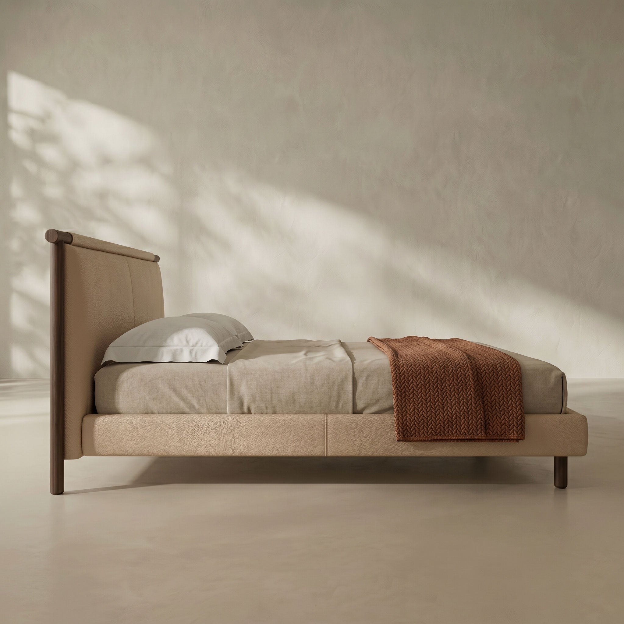 A minimalist single bed with a light beige upholstered frame and dark wood headboard and legs, styled with neutral bedding and a rust-colored throw blanket against a textured beige wall with soft leaf shadows.
