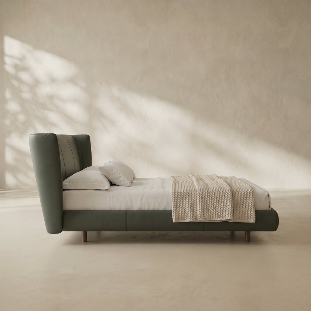A modern upholstered bed with a tall curved headboard and matching footboard, shown in a minimalist room with warm sunlight casting leaf shadows on a textured beige wall and floor.