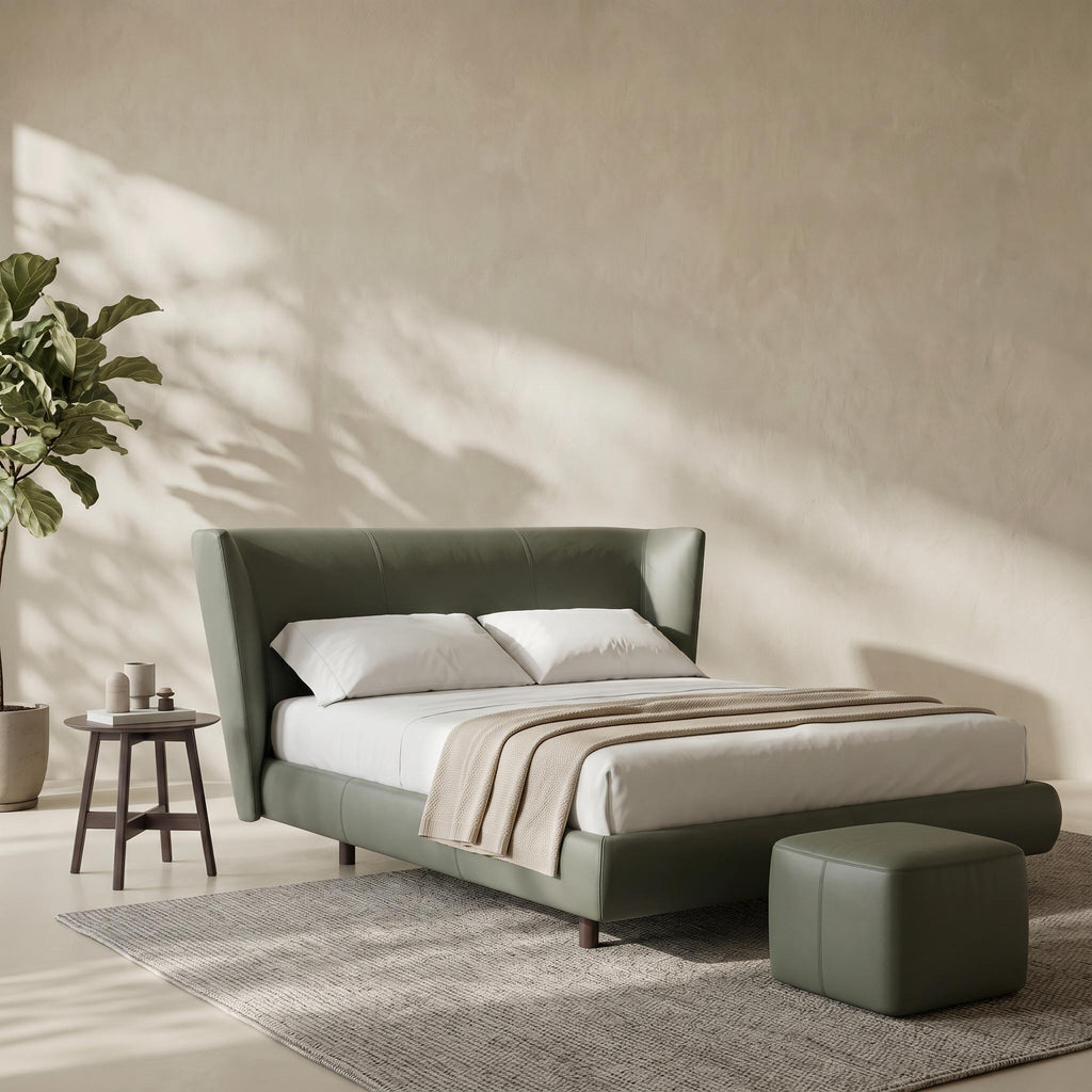 An olive green upholstered bed frame with a tall matching headboard, styled with white bedding and a beige throw blanket, set in a minimalist bedroom with a textured beige wall, a side table, a plant, and a matching green ottoman.