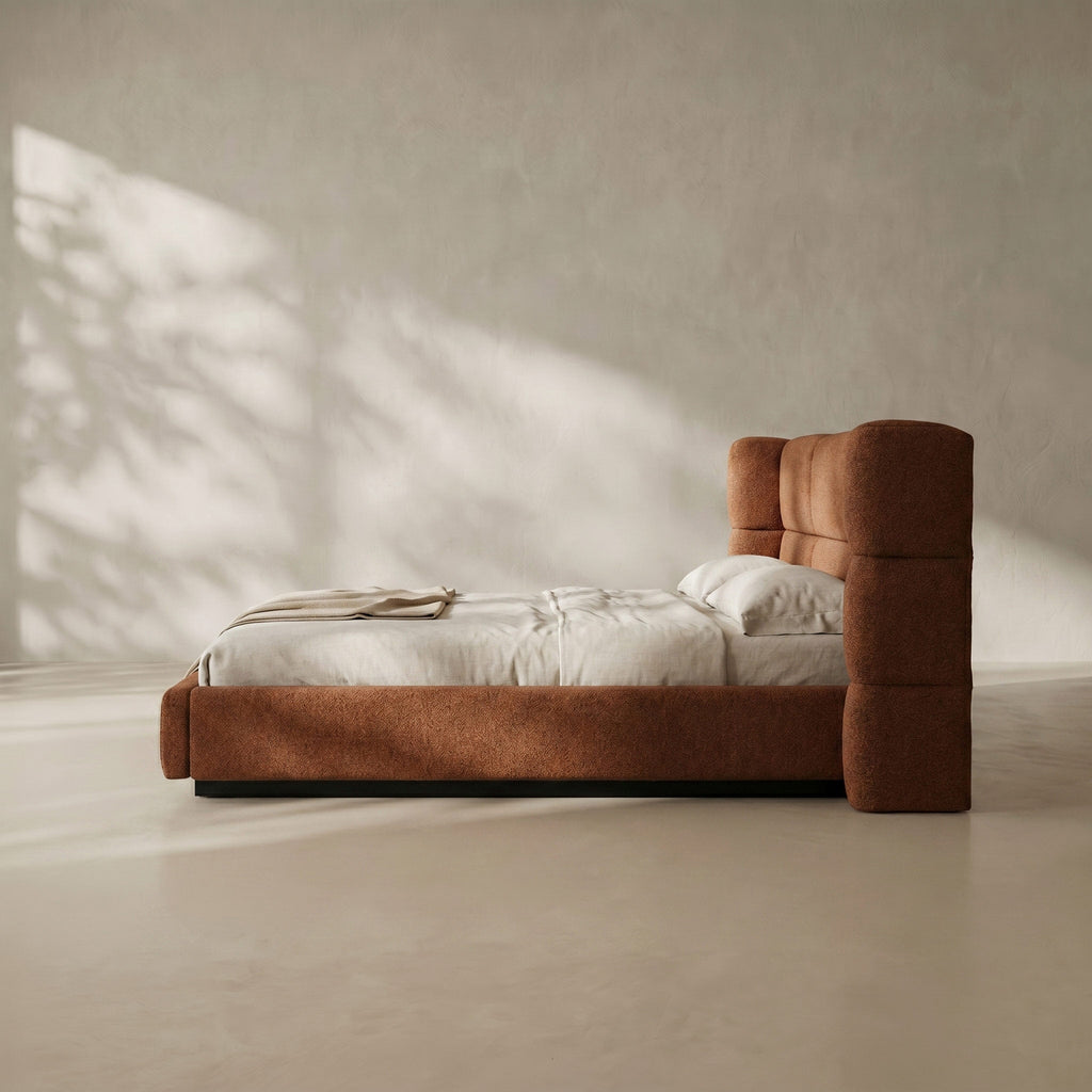 A modern upholstered bed with a tall, blocky tufted headboard and matching footboard in a warm rust-brown fabric, styled with white bedding in a minimalist room with soft leaf shadows.