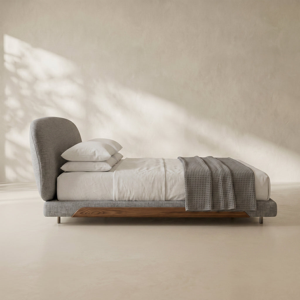 A modern low-profile platform bed with a gray upholstered headboard and footboard, white bedding, two white pillows, and a gray textured throw blanket, set against a warm beige plaster wall with soft window shadow patterns.