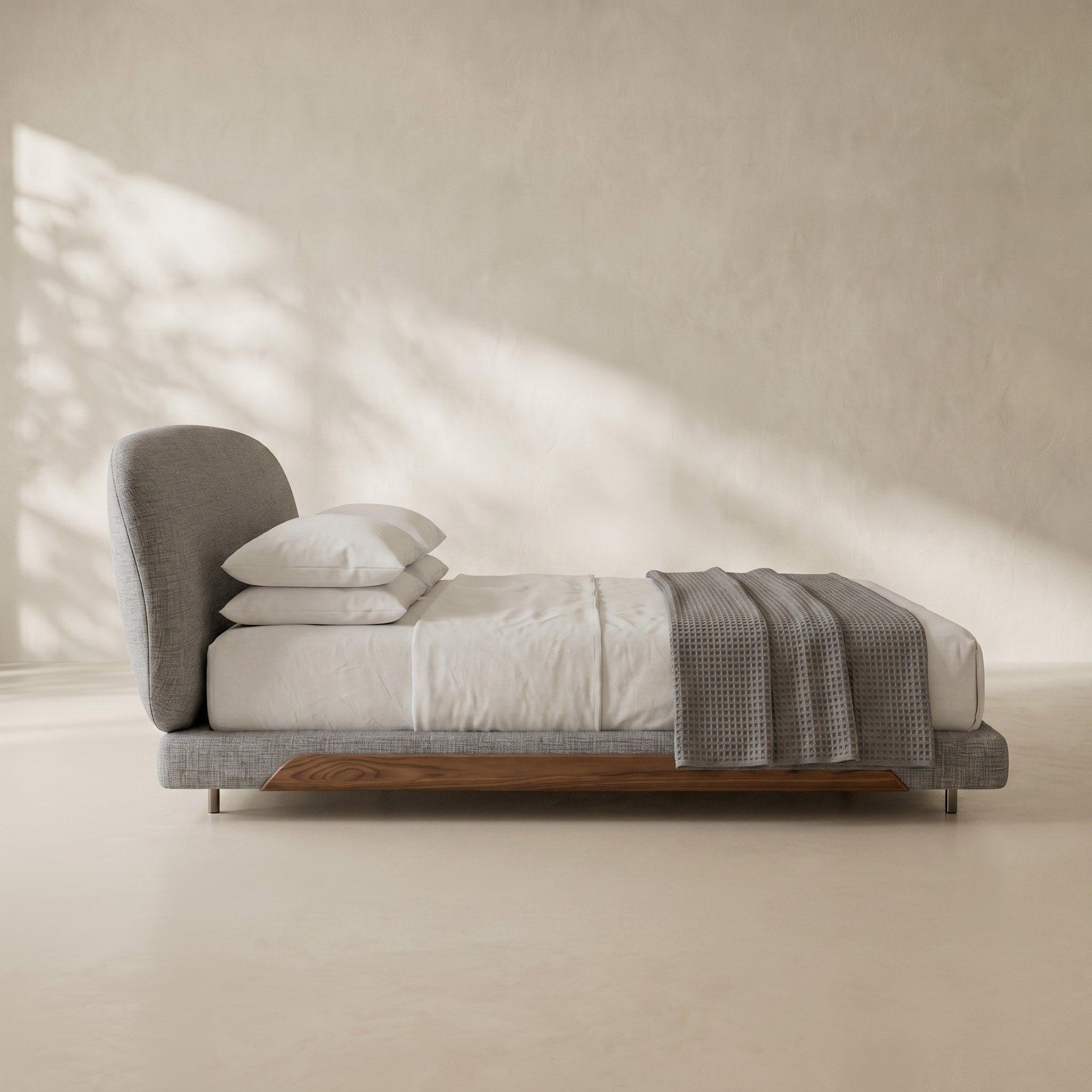 A modern low-profile platform bed with a gray upholstered headboard and footboard, white bedding, two white pillows, and a gray textured throw blanket, set against a warm beige plaster wall with soft window shadow patterns.