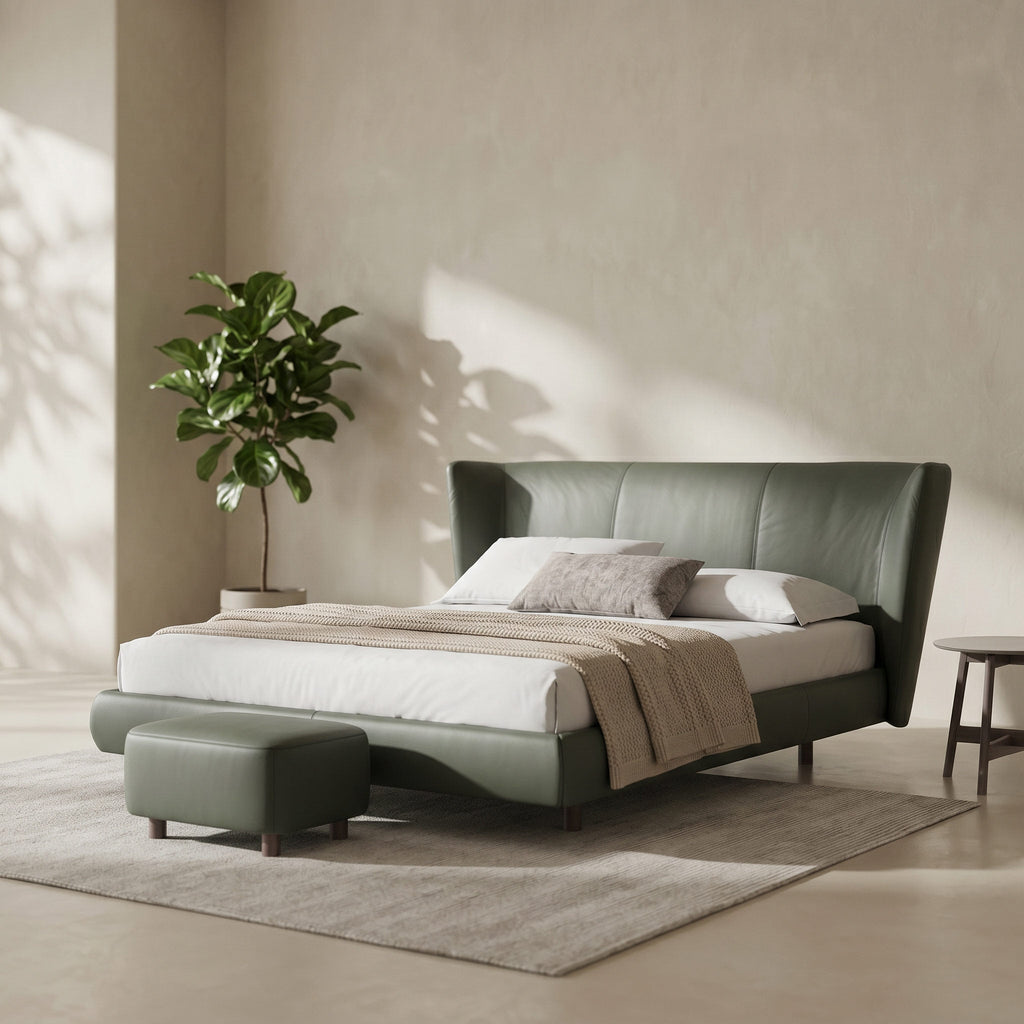 A modern upholstered bed frame in muted sage green, styled in a minimalist bedroom with white bedding, a matching green ottoman, a neutral rug, and a potted plant against a textured beige wall.