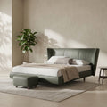 A modern upholstered bed frame in muted sage green, styled in a minimalist bedroom with white bedding, a matching green ottoman, a neutral rug, and a potted plant against a textured beige wall.