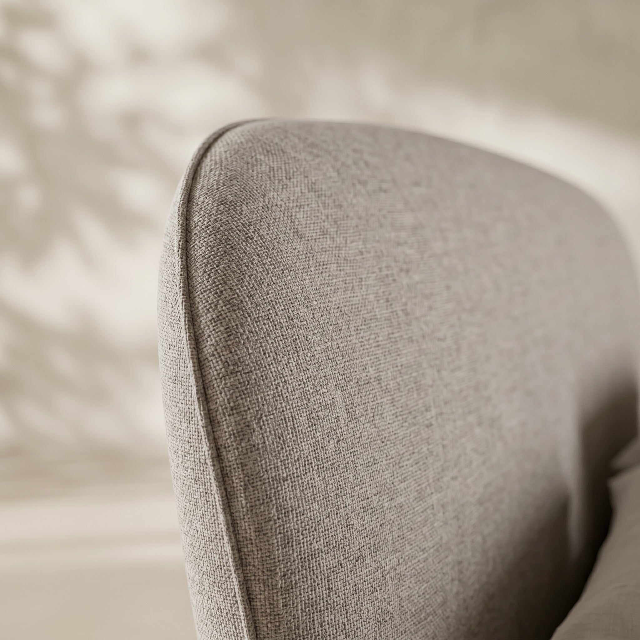 Close-up of a light gray textured fabric upholstered chair or cushion corner with soft warm shadows on a neutral beige background.
