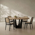 An oval dining table with a light natural wood top and a sculptural black metal pedestal base, surrounded by four matching chairs with black frames, woven cane backs, and light beige seat cushions. The dining set is styled with a bowl of green pears and a glass pitcher on a neutral, textured beige wall with soft leaf shadows.