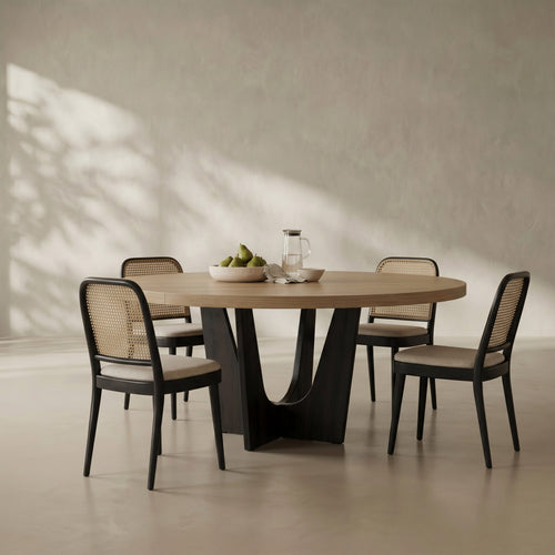 An oval dining table with a light natural wood top and a sculptural black metal pedestal base, surrounded by four matching chairs with black frames, woven cane backs, and light beige seat cushions. The dining set is styled with a bowl of green pears and a glass pitcher on a neutral, textured beige wall with soft leaf shadows.
