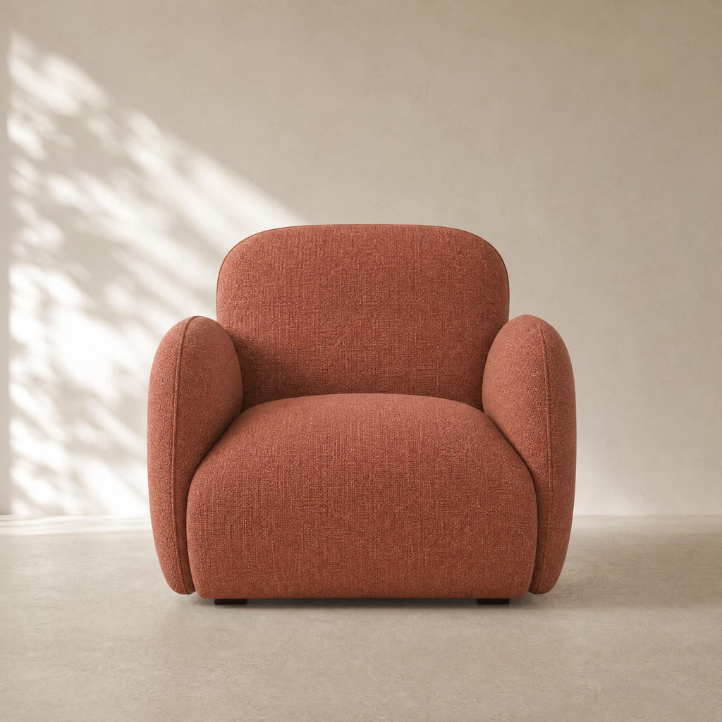 A modern low-profile armchair upholstered in textured terracotta fabric, set against a minimalist beige wall with soft leaf shadows.