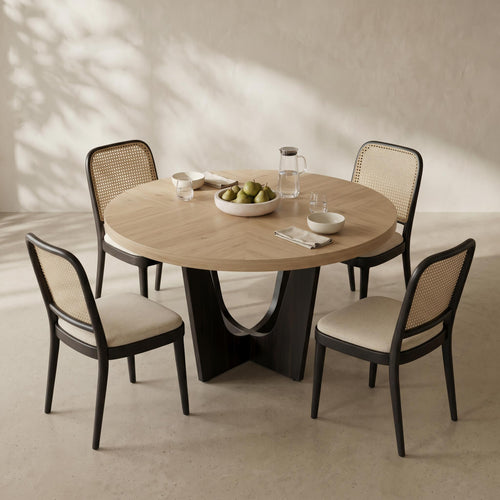 A round light wood dining table with a sculptural black pedestal base, surrounded by four black chairs with woven cane backs and cream upholstered seats, set in a sunlit room with leaf shadows on a textured beige wall.