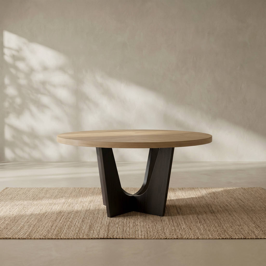 A modern round coffee table with a light natural wood top and a sculptural black metal base, set on a woven jute rug against a textured beige wall with soft leaf shadows.