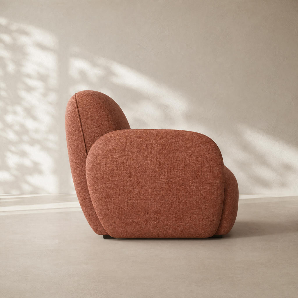 A modern, rounded bubble-style armchair upholstered in textured terracotta fabric, shown from a side profile against a neutral beige wall with soft leaf shadows.