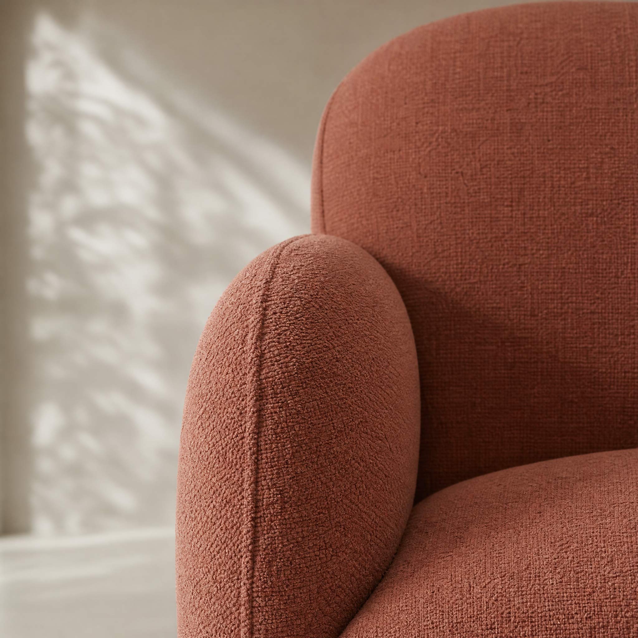 Close-up of a textured upholstered armchair in a warm terracotta or rust color, showing the fabric weave and rounded arm and back cushions against a softly lit neutral interior background.