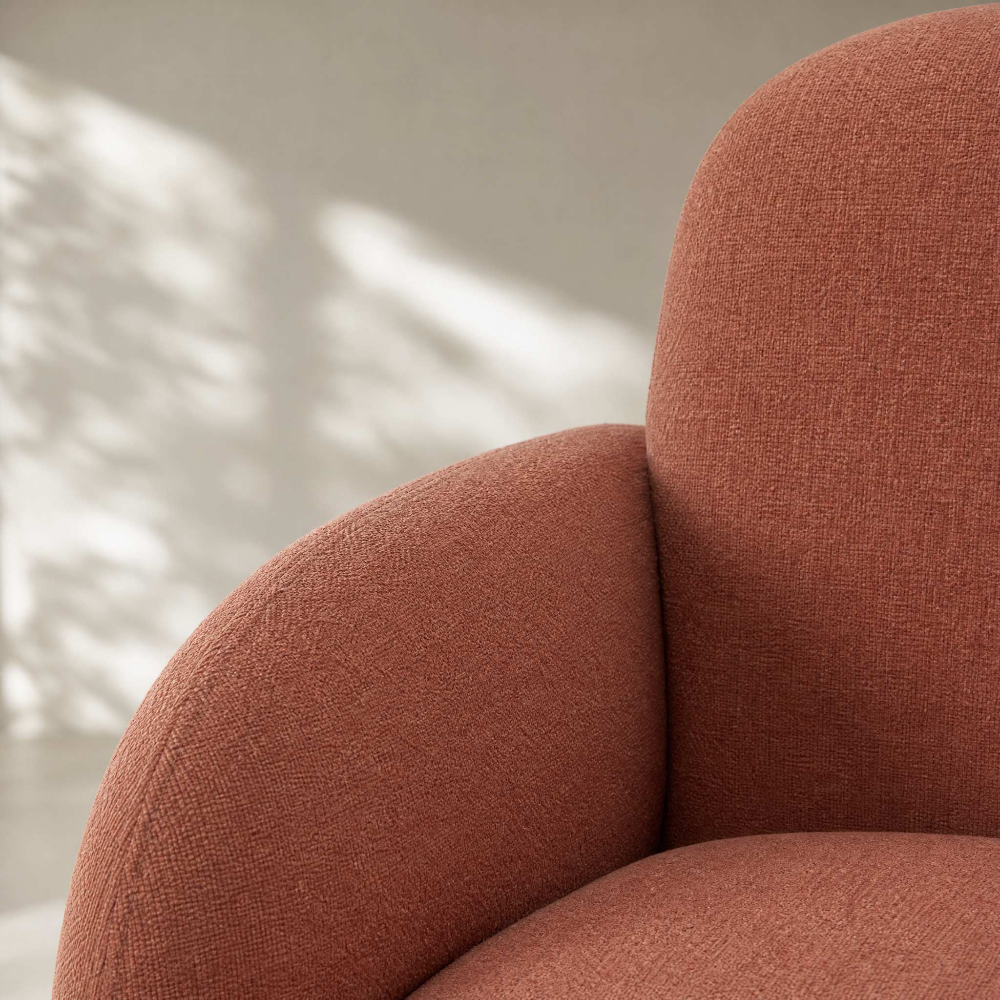 Close-up of a modern upholstered chair in a warm terracotta rust color, showing the textured fabric and curved armrests against a softly lit neutral background.