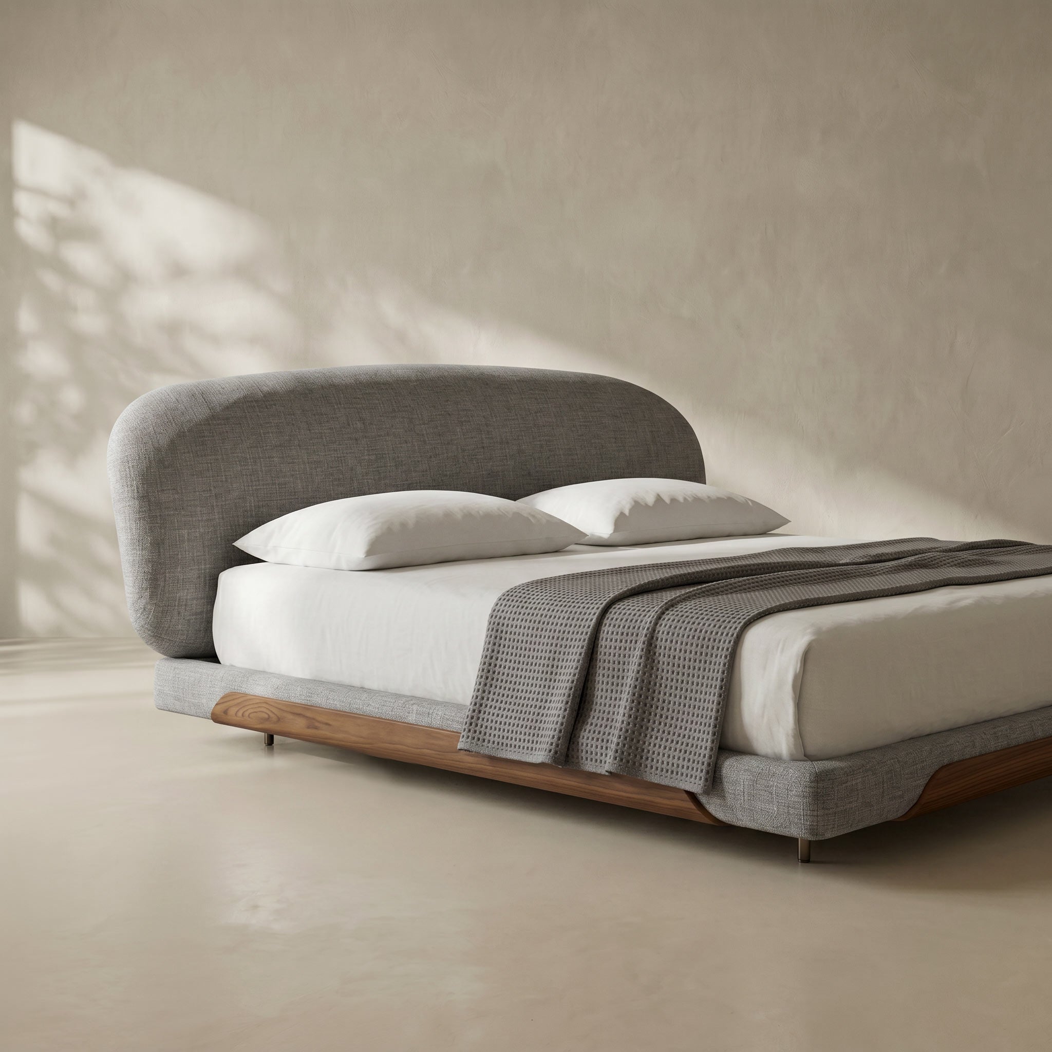 A modern low-profile platform bed with a rounded upholstered headboard, white bedding, and a textured gray throw blanket, set against a warm beige plaster wall with soft leaf shadows.