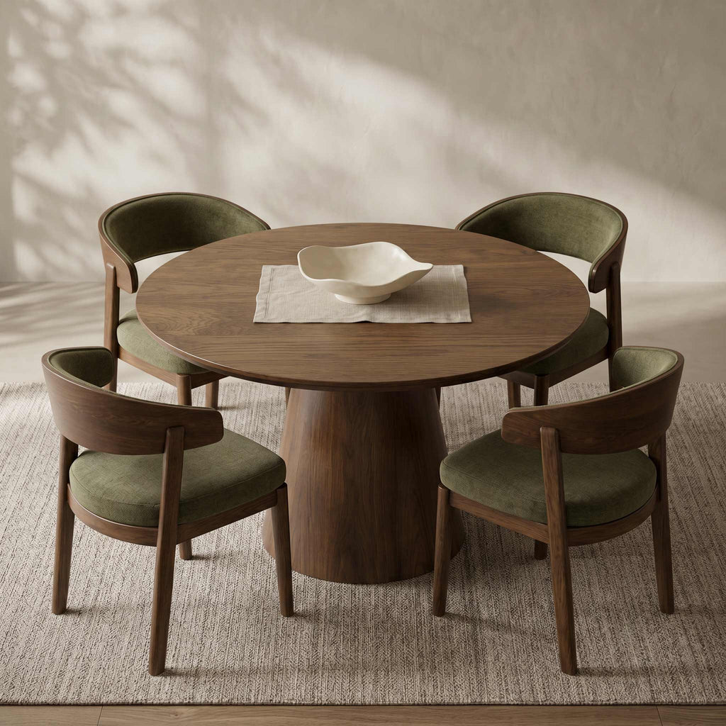 A round wooden dining table with four upholstered chairs in a warm neutral room. The chairs have olive green fabric seats and backs with curved wooden frames.