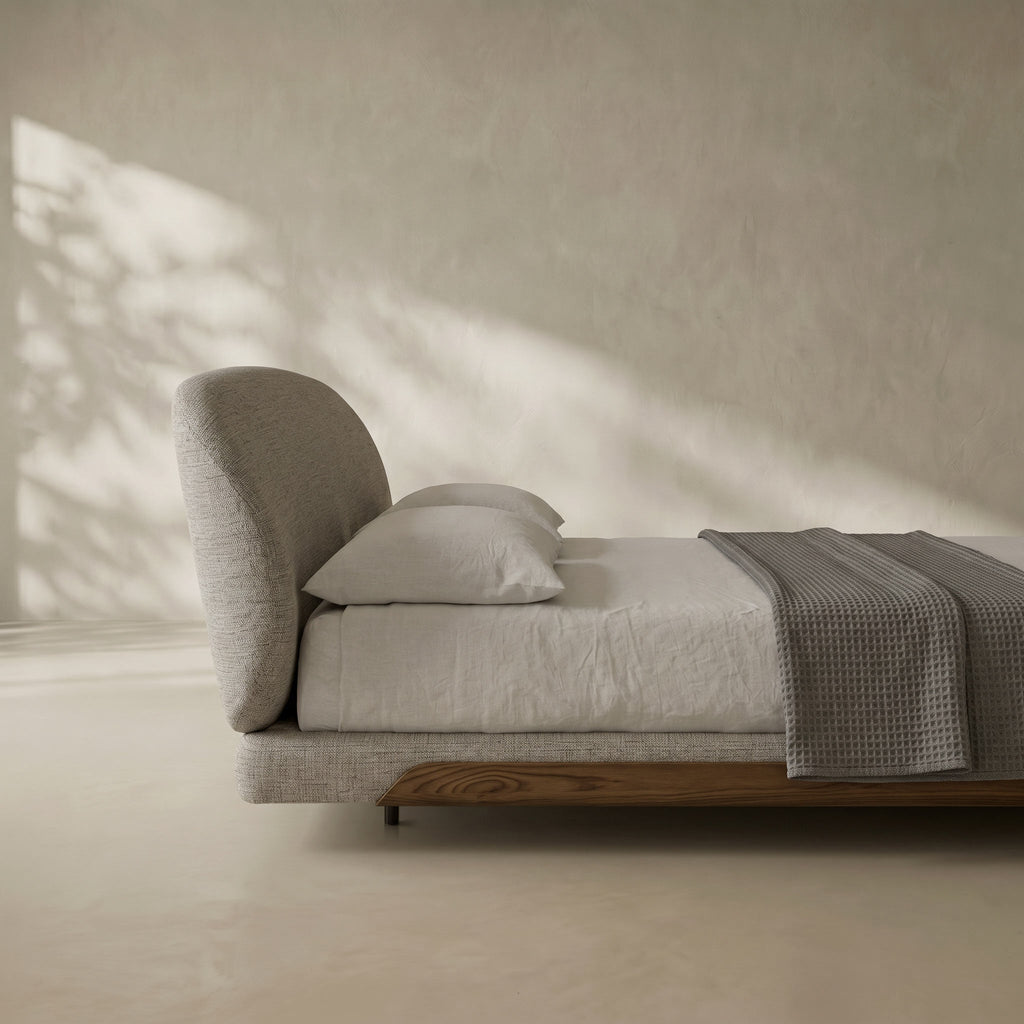 A modern low-profile bed with a tall curved headboard, upholstered in light gray fabric, styled with white linens and a textured gray throw blanket in a minimalist room with warm sunlight and shadows.