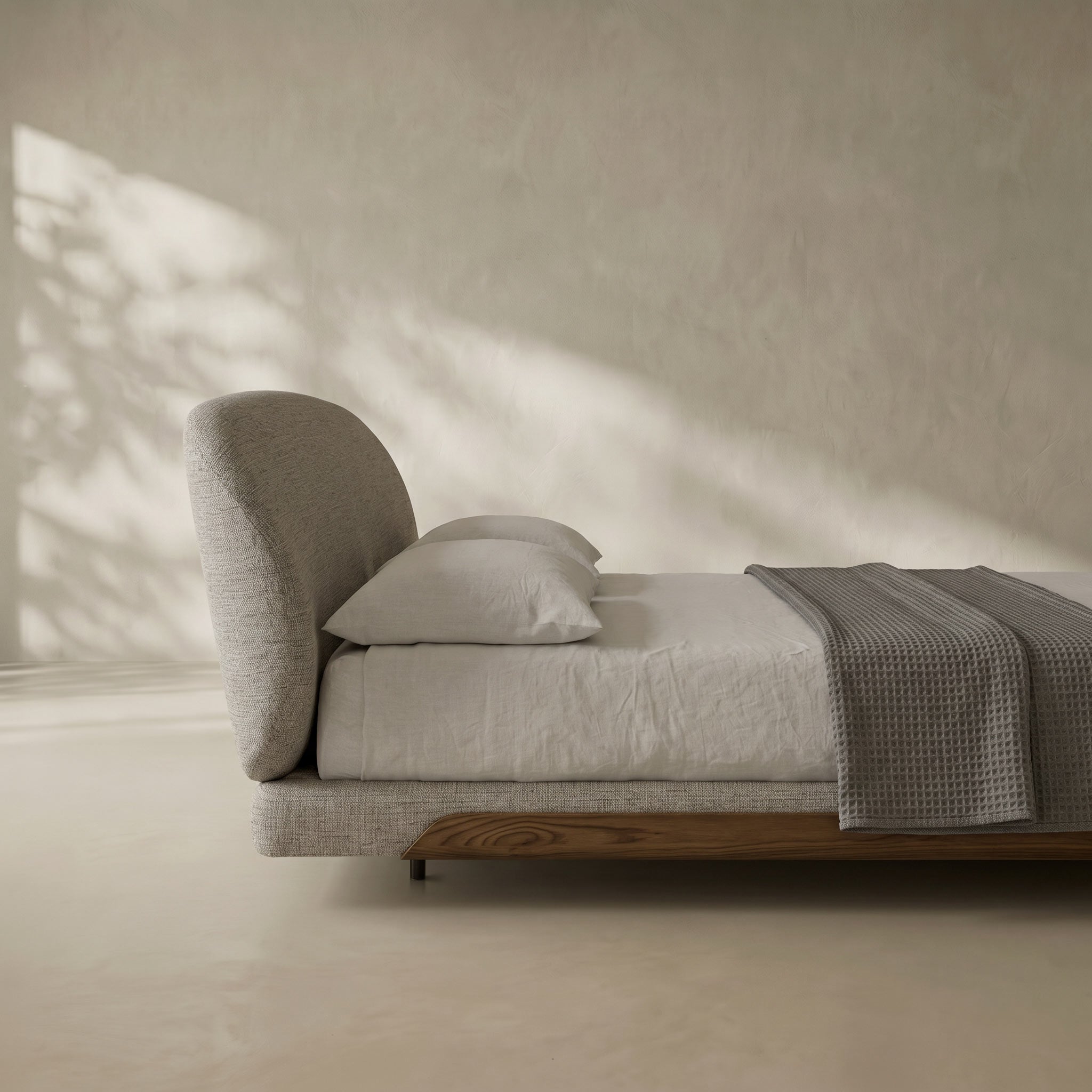 A modern low-profile bed with a tall curved headboard, upholstered in light gray fabric, styled with white linens and a textured gray throw blanket in a minimalist room with warm sunlight and shadows.