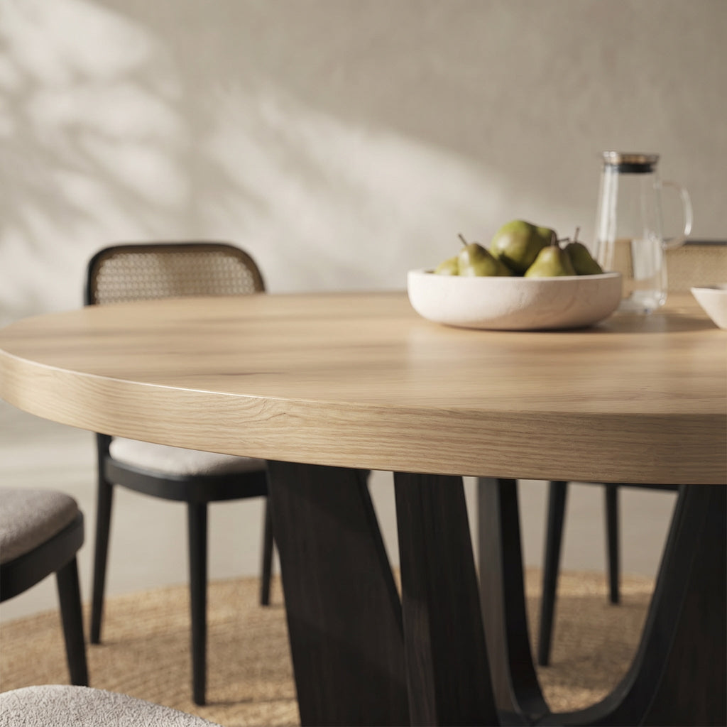 A close-up of a round light oak dining table with dark sculptural legs, styled with a bowl of pears and a glass water carafe in a softly lit room.