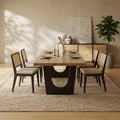 A modern dining room scene featuring a rectangular wooden dining table with a sculptural dark wood base and four matching woven-back chairs with light upholstered seats, set on a textured rug in a warm neutral room.