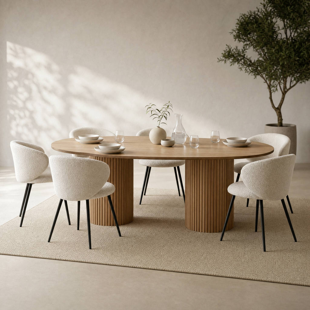 A modern dining set featuring a round light oak wood table with two fluted pedestal legs, surrounded by six upholstered dining chairs with cream bouclé fabric and black metal legs, set on a textured beige rug in a minimalist dining room.