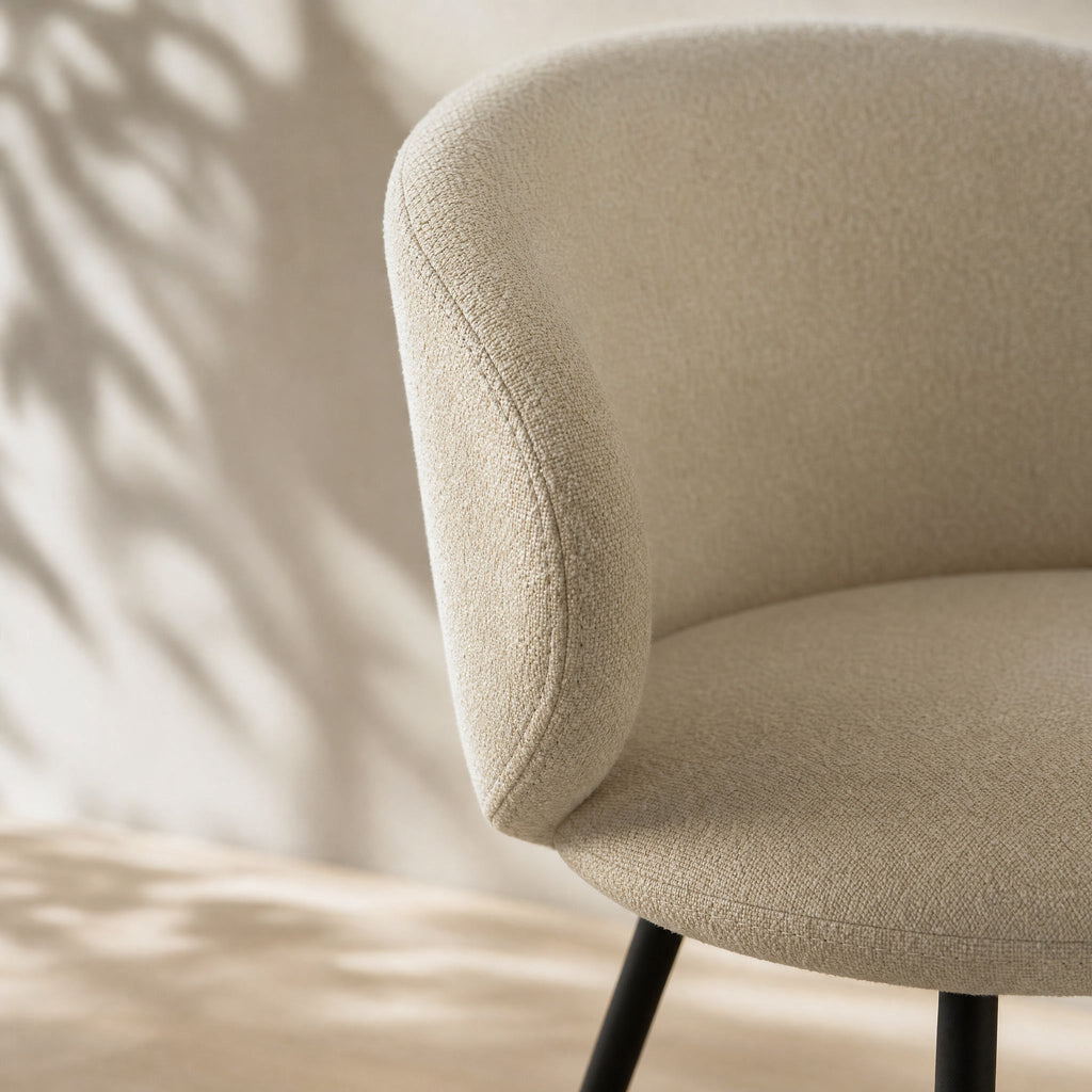 Close-up of a modern upholstered chair in a textured beige fabric, showing the curved backrest and seat with black legs against a softly lit wall with leaf shadows.