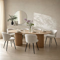 An oval wooden dining table with a fluted pedestal base, surrounded by six upholstered dining chairs with black legs, set in a bright, modern dining room with a round mirror and vase of flowers.