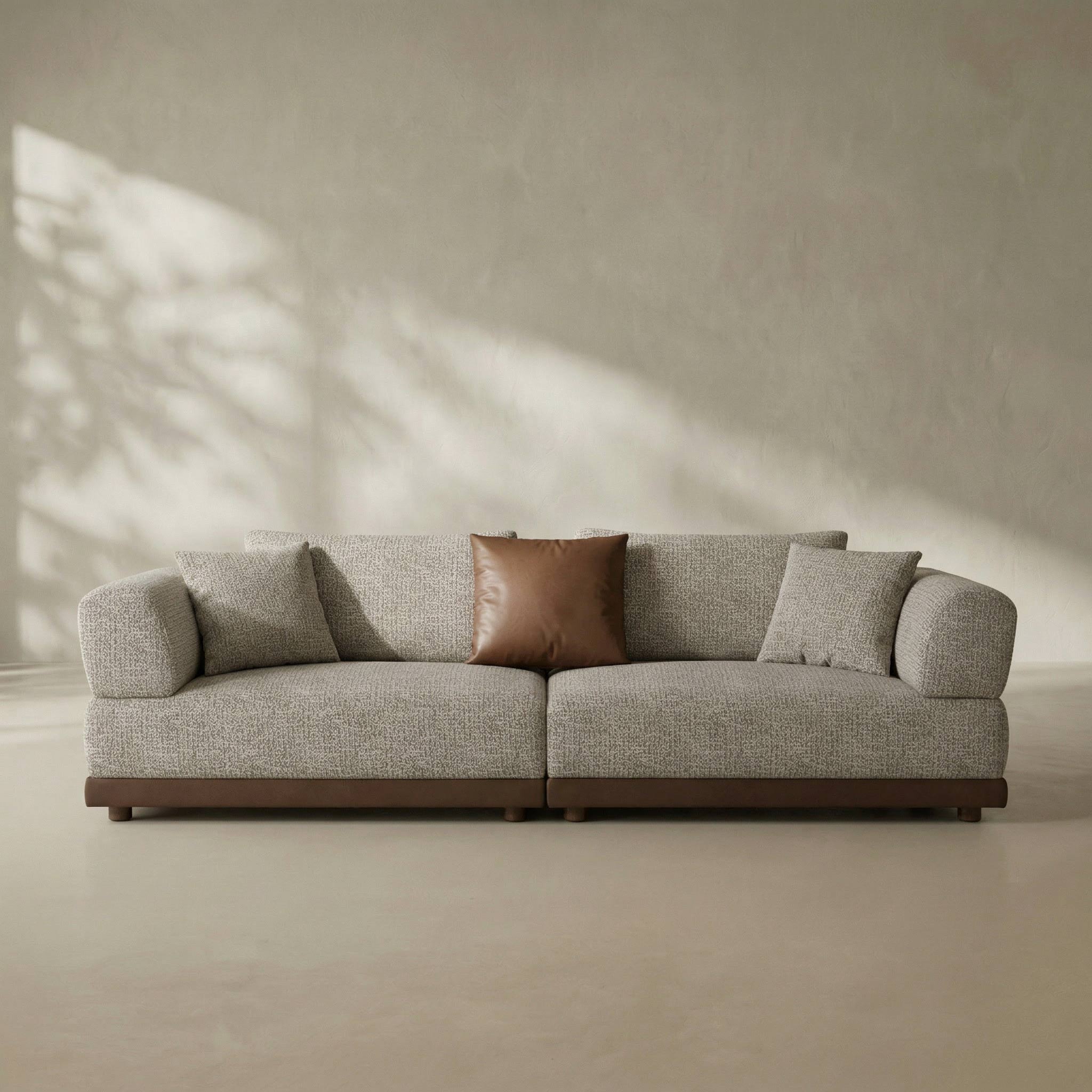 Gray sofa with a brown pillow in a minimalistic room with sunlight casting shadows on the wall.