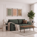 A modern living room featuring a dark green fabric sofa with a brown accent pillow, a beige throw blanket, a wooden coffee table, and three framed textured art prints on a white paneled wall.