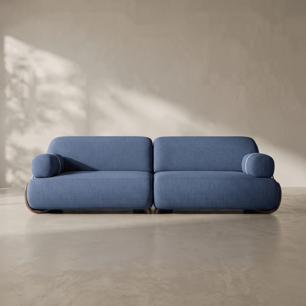 A modern two-seater sofa in textured blue fabric, positioned against a warm beige plaster wall with soft leaf shadows cast across it.