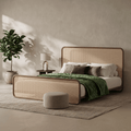 A modern bed with a woven cane headboard and footboard, styled in a minimalist bedroom with a green throw blanket, neutral pillows, a small side table with a lamp, a potted plant, and a round ottoman.