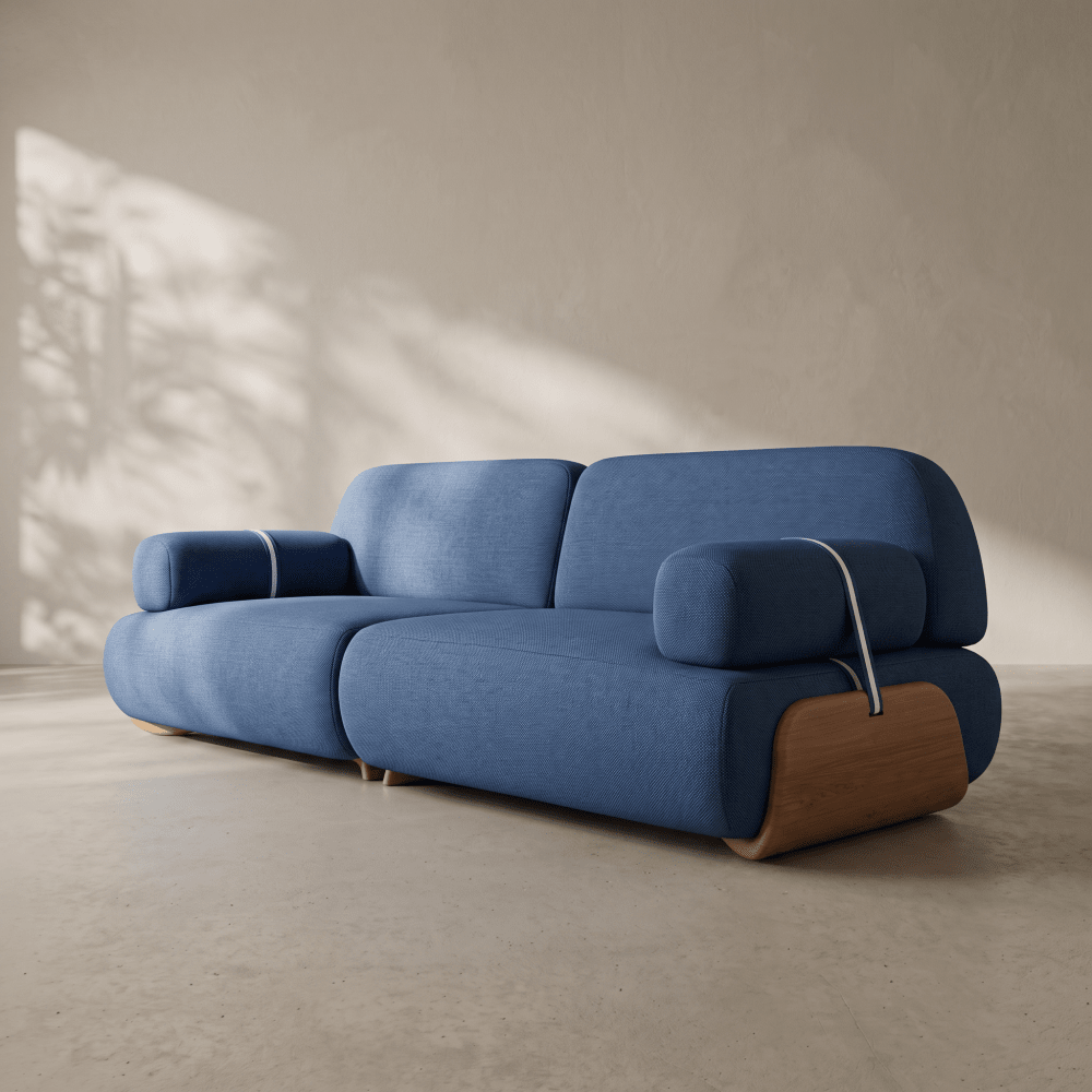 A modern blue upholstered sofa with rounded blocky cushions and exposed wood side supports, photographed in a minimalist interior with warm sunlight and shadows on a beige wall and floor.