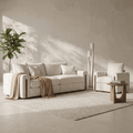 A modern living room setup featuring a cream-colored sofa and matching armchair on a textured light rug, with a small wooden side table, a tall potted plant, and a minimalist floor lamp against a textured beige wall.