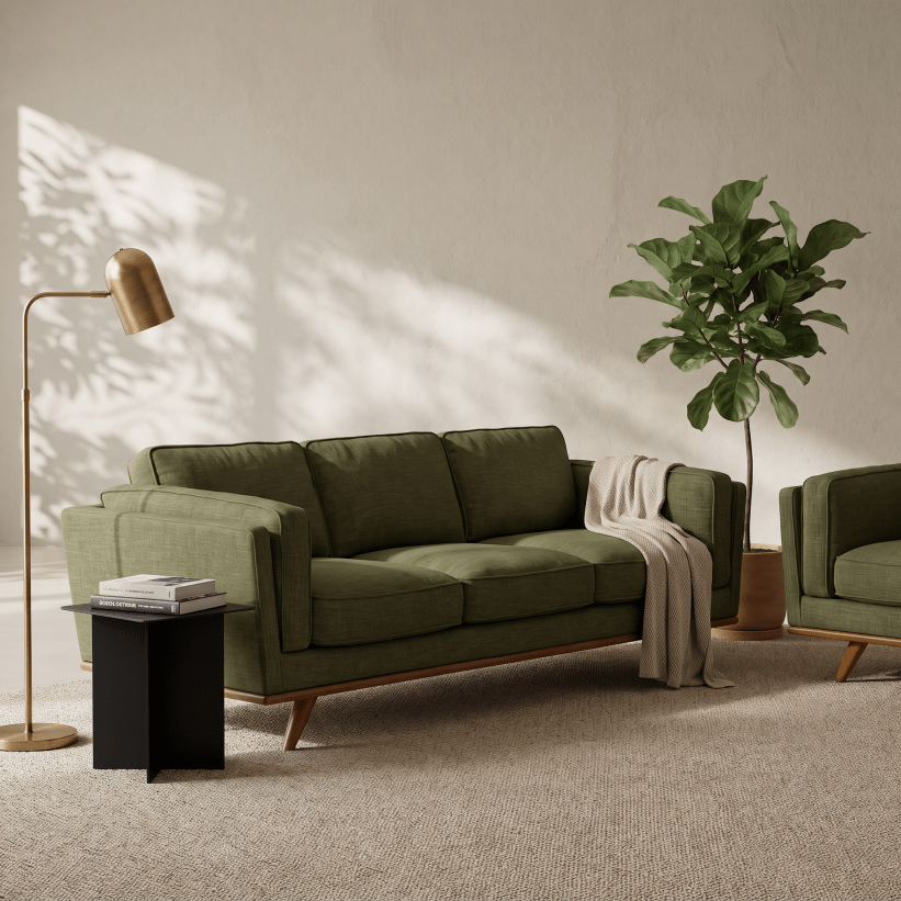 An olive green fabric sofa with a mid-century modern design, featuring tapered wooden legs and a draped cream throw blanket, set in a styled living room with a floor lamp, side table, and potted plant.