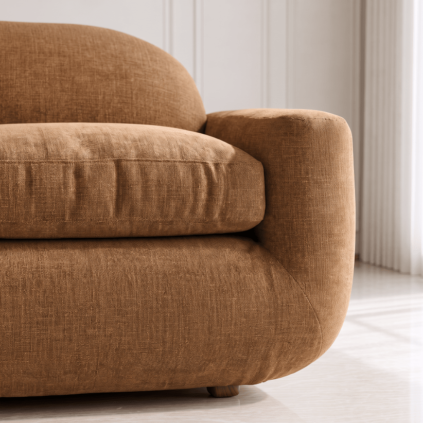 Close-up of a modern armchair in textured tan-brown fabric, showing the seat cushion, rounded armrest, and upholstered base in a bright interior setting.