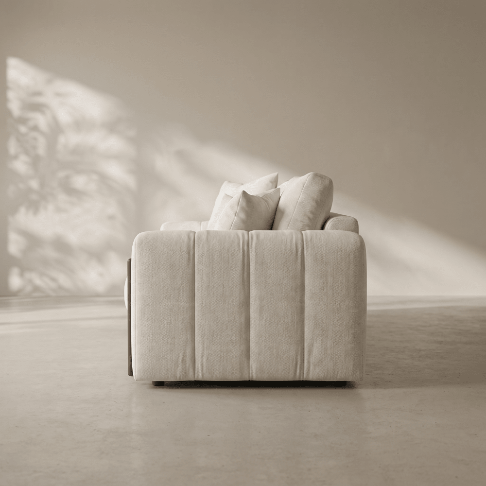 A modern low-profile sofa in a light beige textured fabric, featuring vertical channel tufting and plush matching back cushions, set in a minimalist room with soft natural light casting leaf shadows on the wall.