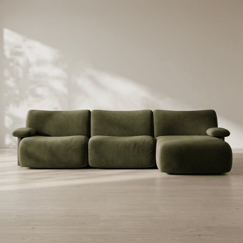 A modern three-seat sofa upholstered in olive green velvet, positioned on a light wood floor against a neutral beige wall with soft leaf shadows.