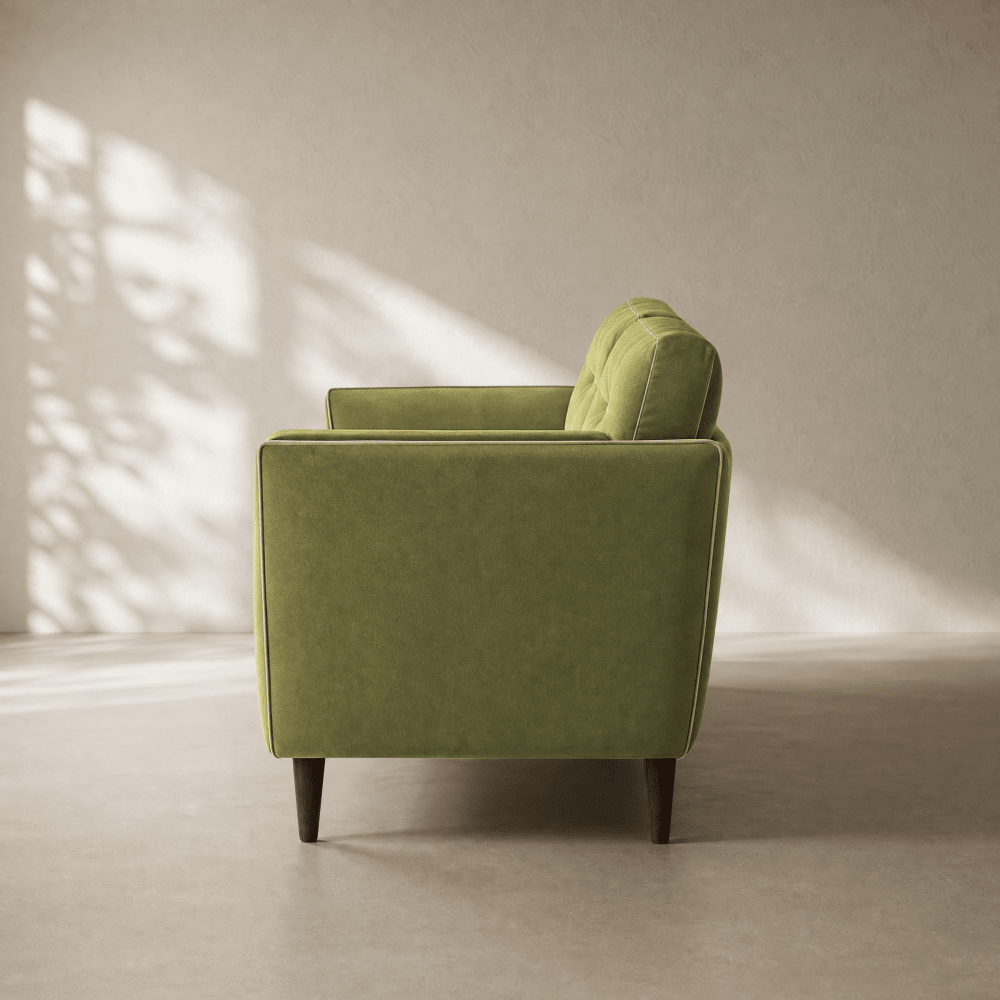 A single upholstered armchair in a muted sage green velvet fabric, shown from a side angle in a minimalist room with soft window shadow patterns on the wall and floor.