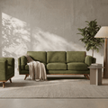 An olive green three-seater sofa with mid-century wooden legs, styled in a modern living room with a beige throw blanket, a matching armchair, a side table, and a potted plant against a textured concrete wall.