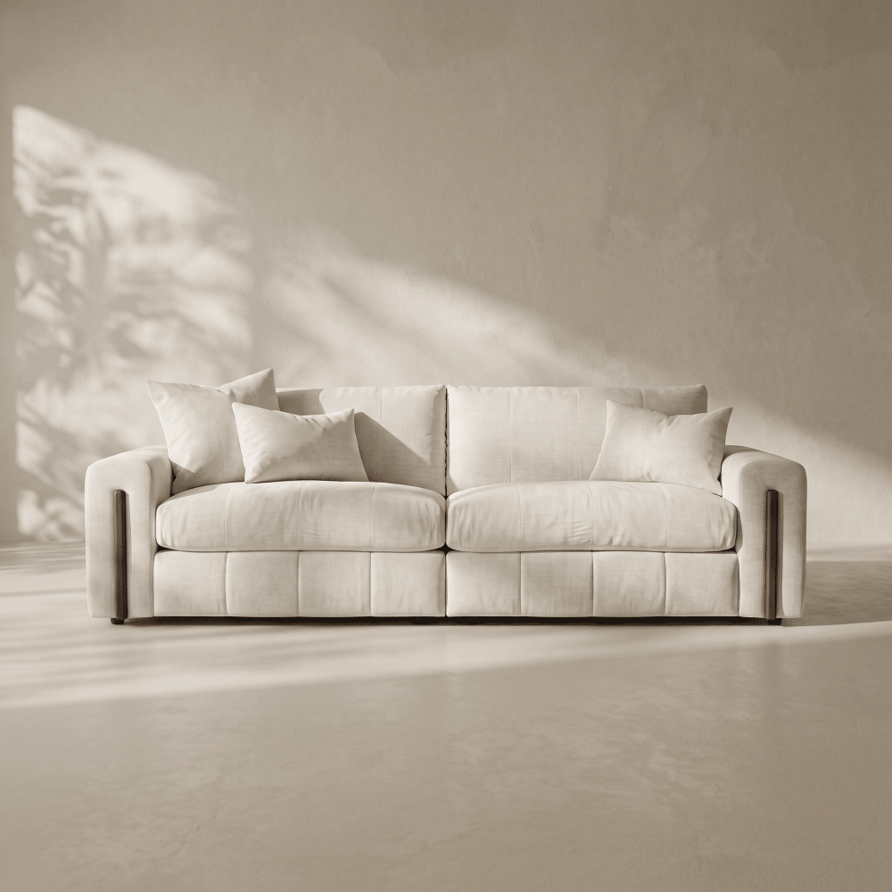 A modern two-seater sofa upholstered in a light cream fabric, styled with matching throw pillows, set against a minimalist beige wall with soft leaf shadows cast across it.
