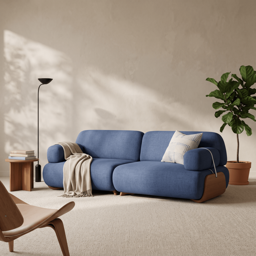 A blue upholstered sofa in a styled living room setting, with a beige throw blanket draped over one arm, a patterned accent pillow, a wooden side table with books and a floor lamp, and a potted fiddle-leaf fig plant against a textured beige wall.