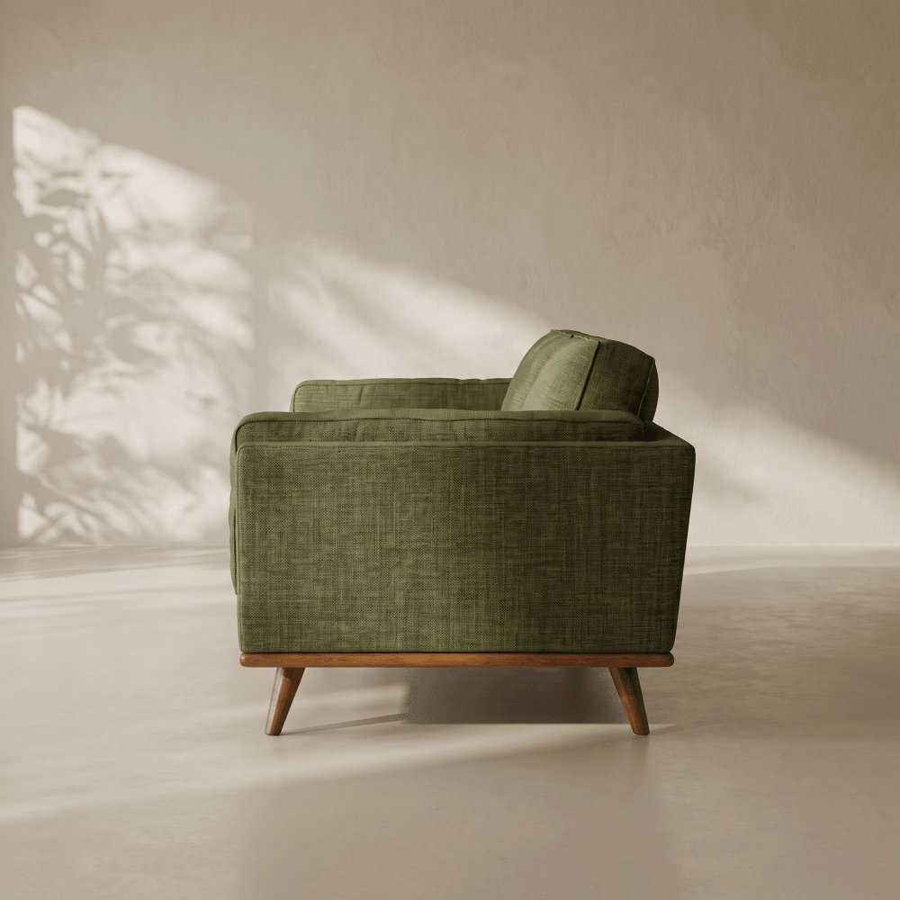 An olive green upholstered armchair with a mid-century wooden base and tapered legs, shown in profile against a warm beige wall with soft leaf shadows.