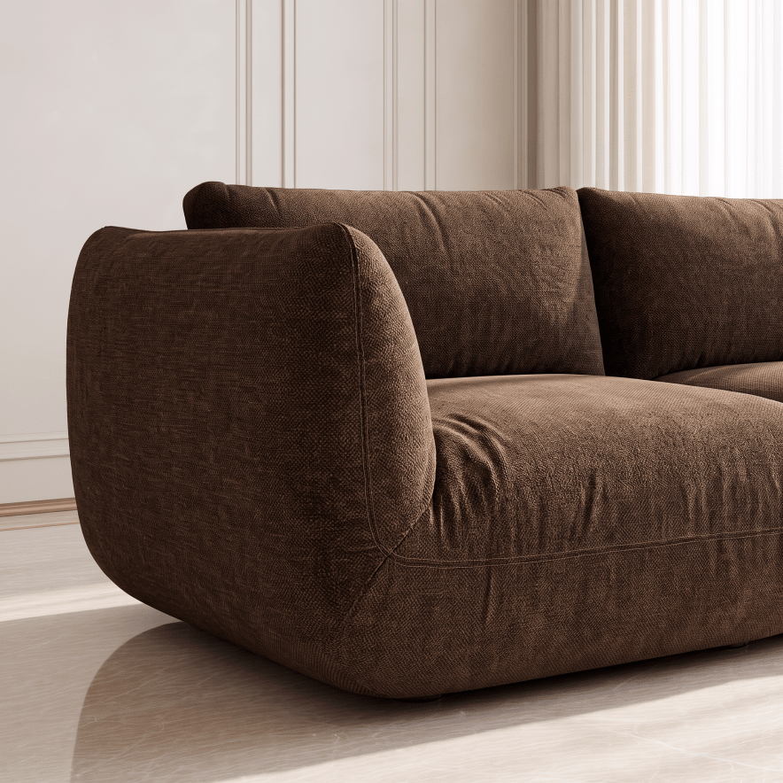 A close-up of a modern brown upholstered sofa in a bright, minimalist room with cream walls and light wood flooring.