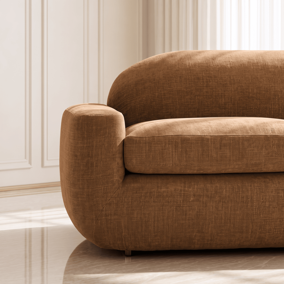 A close-up of a modern upholstered sofa corner in a warm camel brown fabric, set against a softly lit cream paneled wall and light-colored floor.